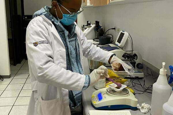 A student wearing a University of Notre Dame lab coat and a face mask uses a small spatula to add a substance to a scale.  A notebook and various lab materials rest on the counter.