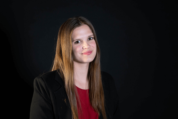A headshot of a young woman with long brown hair against a black background. She wears a black blazer and a dark red shirt. She is smiling slightly and looking off to the right side of the frame.