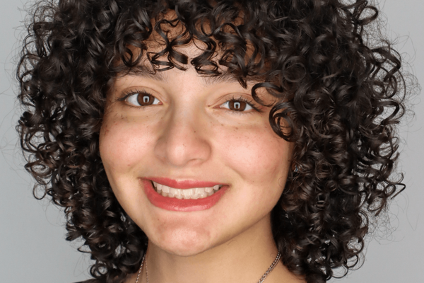 Headshot of a young person with tight, dark curly hair, brown eyes, and a broad smile against a plain gray background.