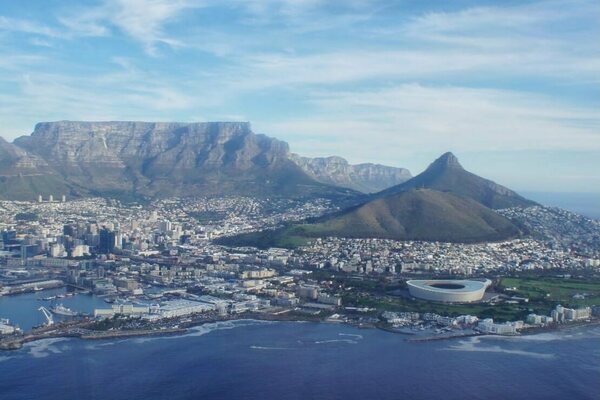 Aerial view of Cape Town, South Africa, with Table Mountain in the background, Lion's Head to the right, and the city sprawling towards the coastline.  The ocean is a deep blue, and a large stadium is visible near the shore.