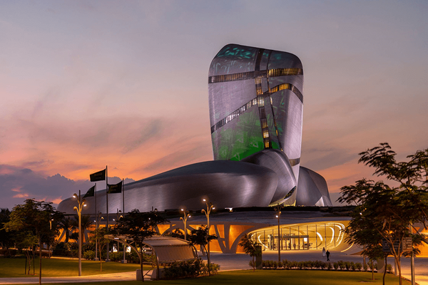 The Museum of the Future in Dubai, UAE, at dusk. The unique silver and green, organically shaped building rises against a vibrant orange and purple sunset. Green light illuminates sections of the facade.  Flags fly in the landscaped area in front of the building. Two figures walk towards the entrance.