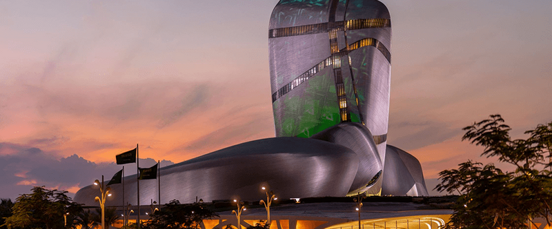 The Museum of the Future in Dubai, UAE, at dusk. The unique silver and green, organically shaped building rises against a vibrant orange and purple sunset. Green light illuminates sections of the facade.  Flags fly in the landscaped area in front of the building. Two figures walk towards the entrance.