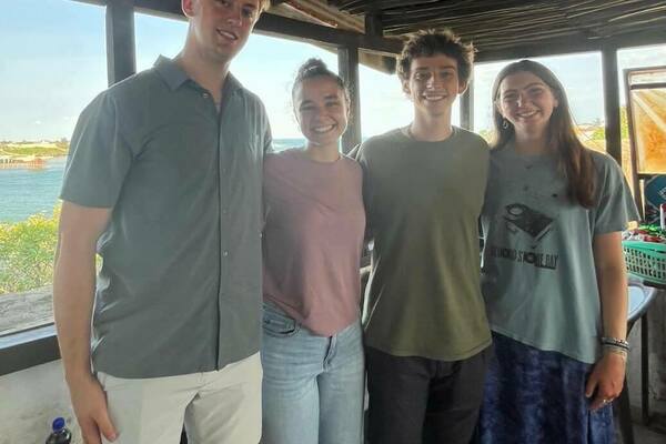 Four students smile for a photo inside a thatched-roof structure with a view of water in the background.