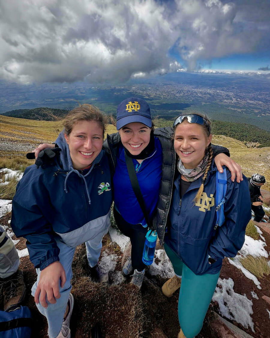 2024-25 Study Abroad Photo Contest ND Irish Abroad Winner Grace Leeson. Three smiling students wearing Notre Dame apparel pose together on a snowy mountaintop overlooking a valley and distant city under a cloudy sky.