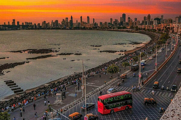 Aerial view of a bustling coastal road in Mumbai, India, at sunset. A red double-decker bus navigates an intersection, surrounded by other vehicles and pedestrians. The vibrant orange and pink sunset illuminates the cityscape in the background.