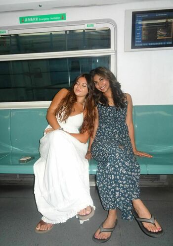 Two young women sit on a green subway bench. The left woman with long brown hair wears a white dress, smiling with closed eyes. The right woman with dark hair wears a dark blue floral dress, smiling at the camera.