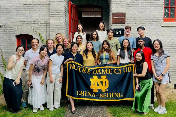 A diverse group of 22 smiling Notre Dame Club members hold a blue and gold "NOTRE DAME CLUB CHINA - BEIJING" banner in front of a gray brick building with red window frames.
