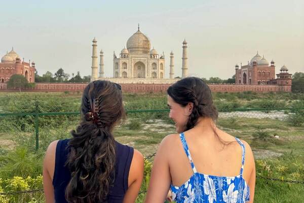 Two women view the white marble Taj Mahal from behind a fence. The woman on the left has dark curly hair in a clip and wears a dark top. The woman on the right has braided hair and wears a blue and white floral dress, smiling slightly. Red sandstone buildings flank the monument.