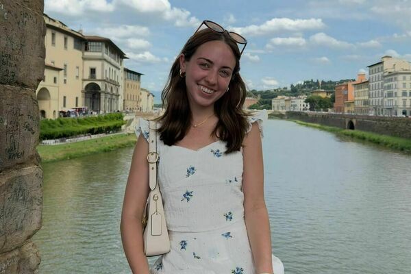 A young woman in a white sundress with blue floral patterns smiles brightly, sitting on a stone wall overlooking a river lined with historic European buildings under a cloudy sky.