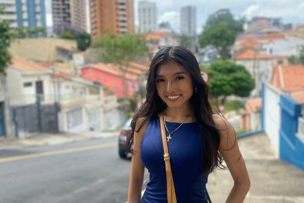 Smiling young woman in blue top & jeans, gold cross necklace, holding a woven bag, stands on a hilly urban street. Background shows diverse buildings with colorful walls & tiled roofs, and taller city structures under a cloudy sky.