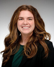Ellen Budell. A smiling woman with long, wavy brown hair, wearing a dark green top and black blazer against a light grey background.
