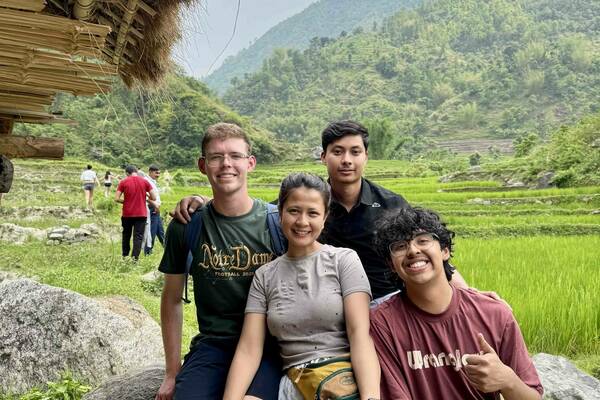 Four smiling young people sit on rocks. One wears a green "Notre Dame Football 2025" shirt. Behind them are vibrant green rice paddies, lush mountains, and a cloudy sky. The person on the right, wearing a maroon shirt, gives a thumbs-up. A thatched roof is visible on the left.