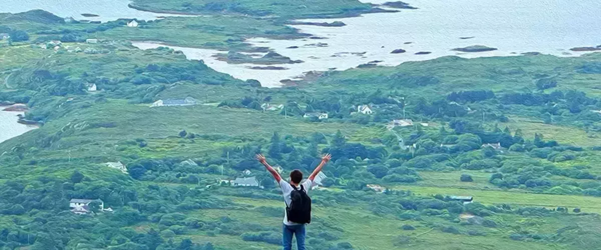 Person in a white shirt and blue jeans with a black backpack stands on a green cliff, arms raised, overlooking a sprawling green landscape with white houses and islands in light blue water.