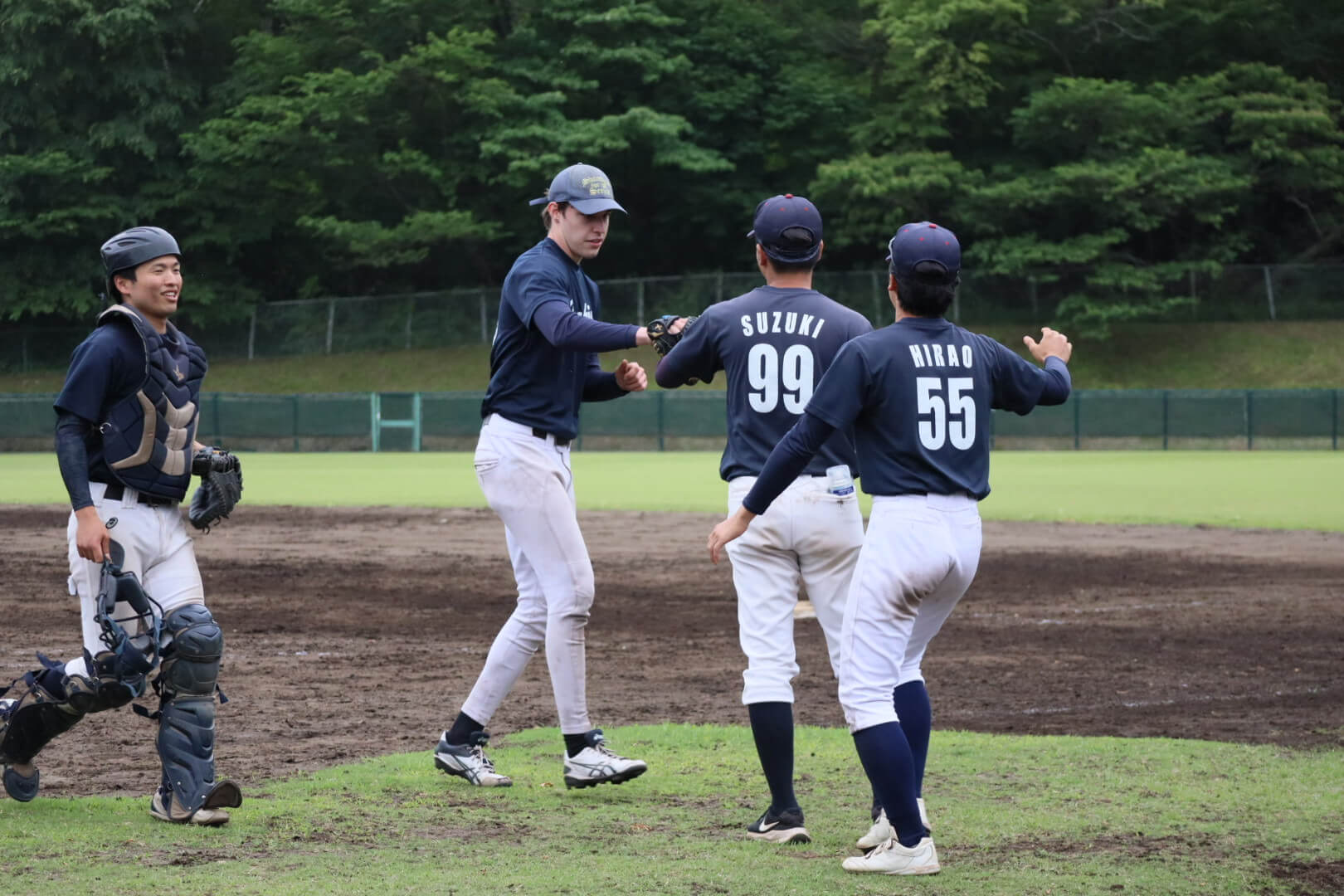 A smiling catcher watches two baseball players in navy uniforms shake hands on a dirt and grass field.
