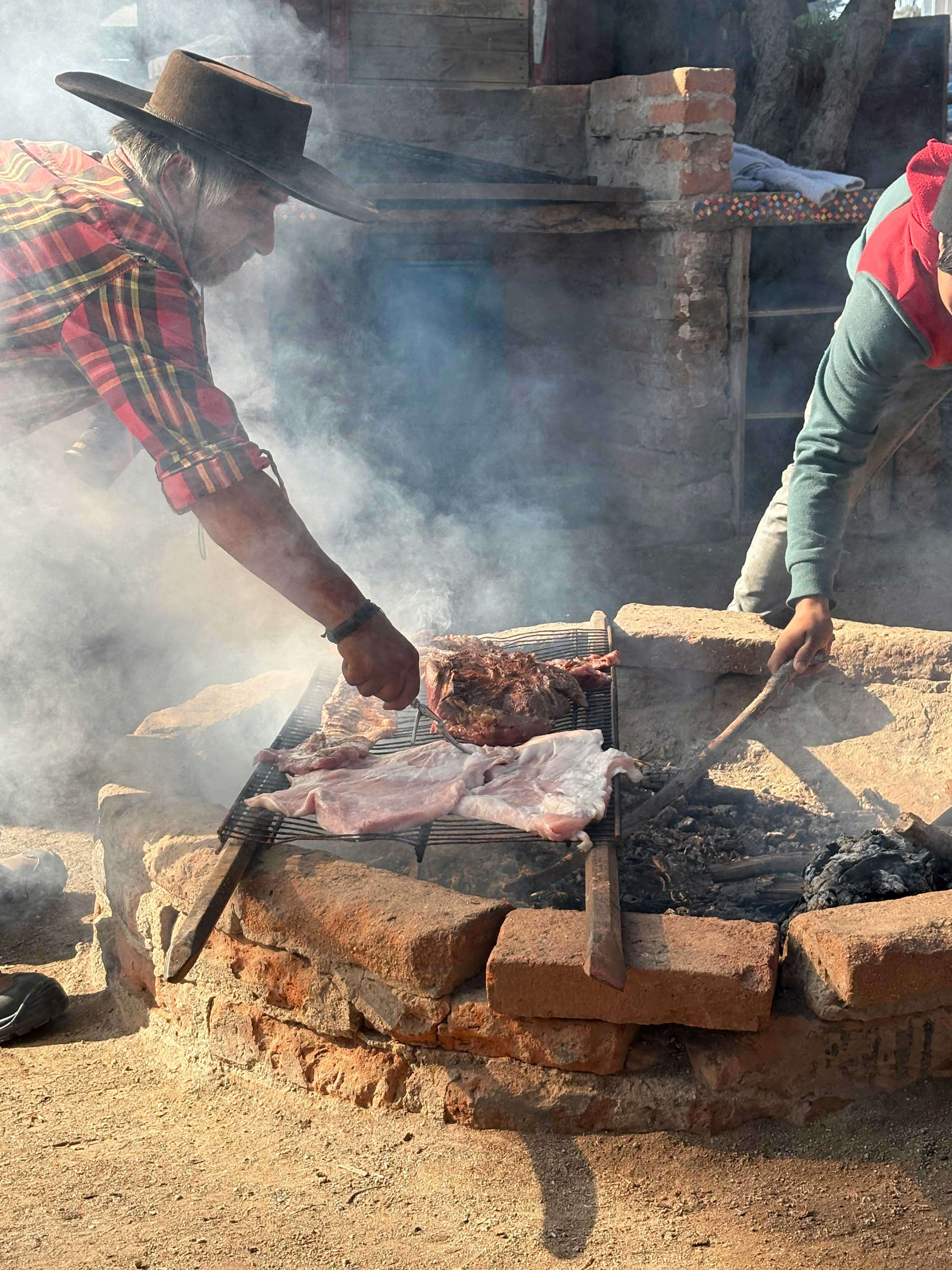 Older man in hat and plaid shirt turns meat on a smoky outdoor brick grill, another person stirs coals nearby.