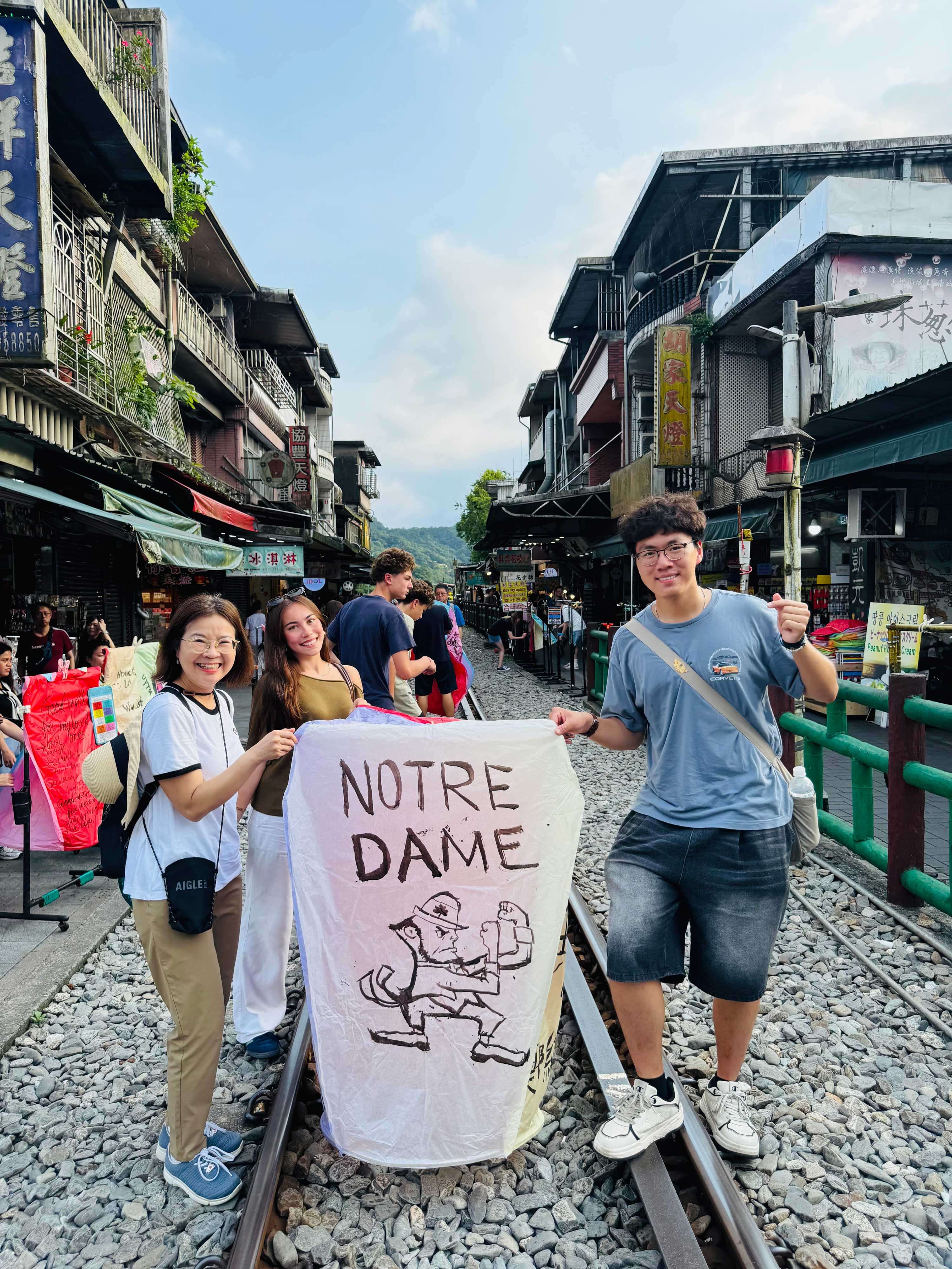 Three smiling people hold a white sky lantern with 'NOTRE DAME' and Leprechaun art, standing on train tracks.