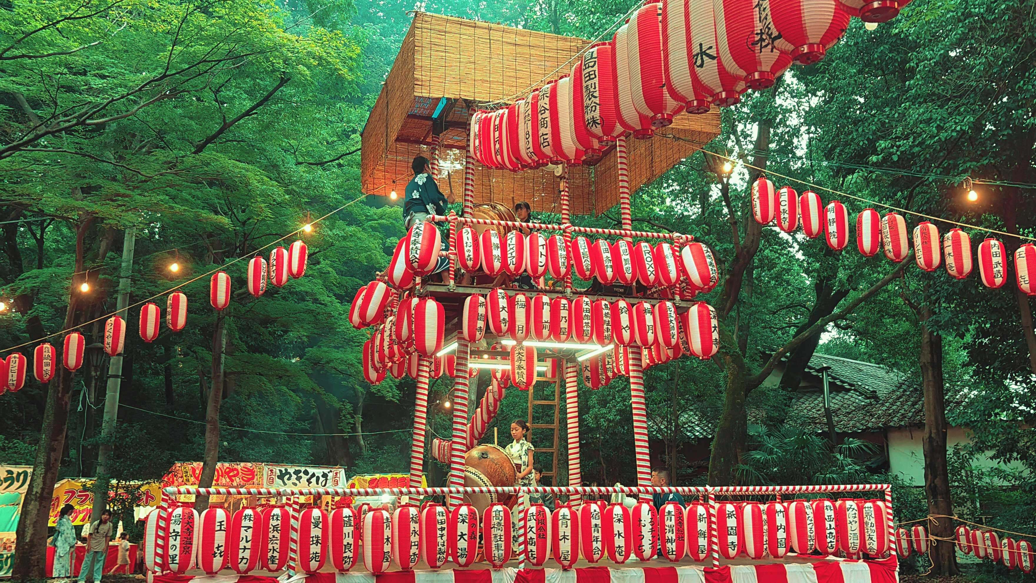 Red and white lanterns adorn a multi-tiered festival stage where people play taiko drums, set amidst lush green trees.