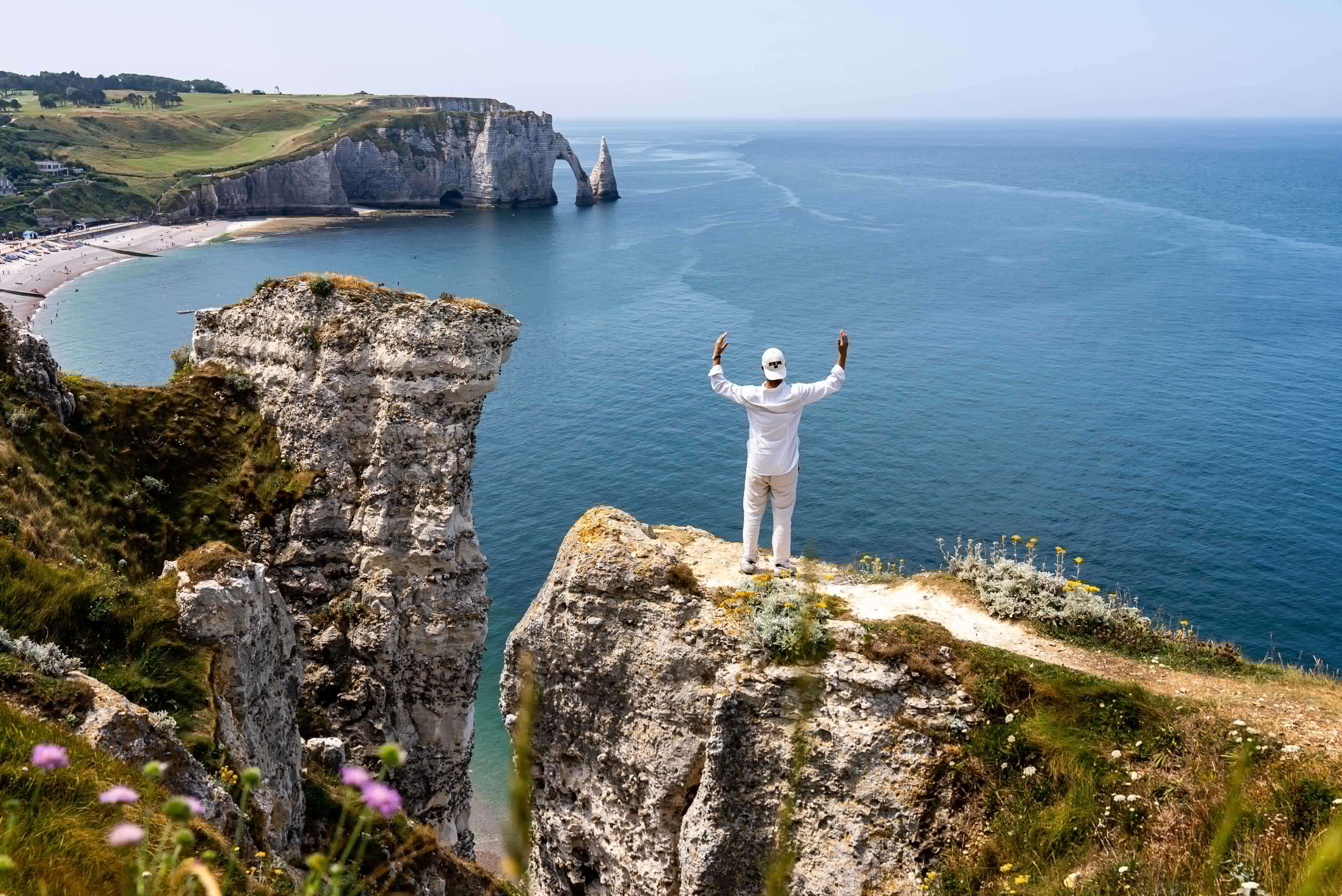 Man in white with raised arms on Etretat cliff. Chalk arches, including Porte d'Aval and Aiguille, and blue sea in background.