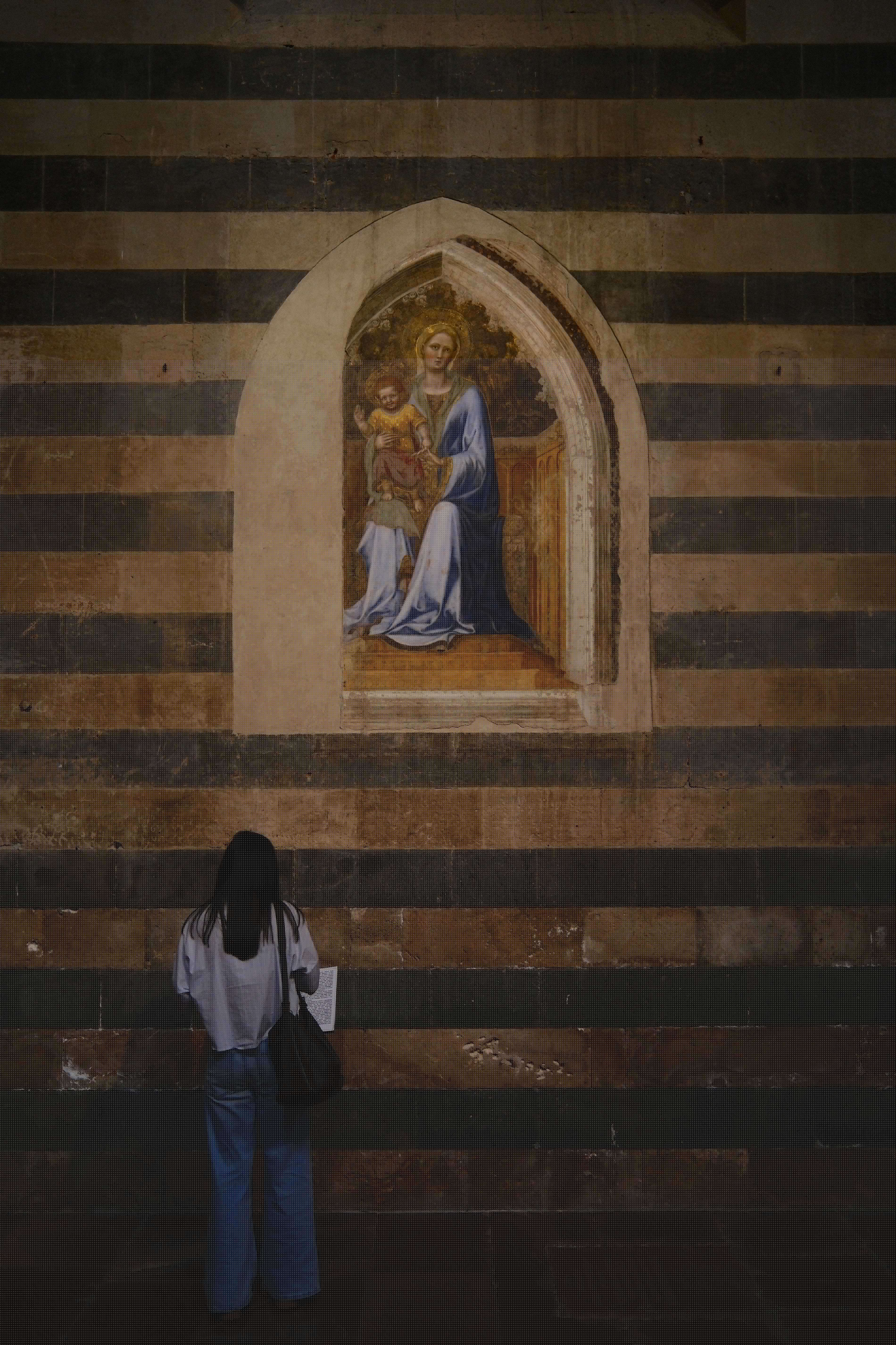 Woman views Madonna and Child painting in an arched niche on a wall with horizontal black and brown stripes.