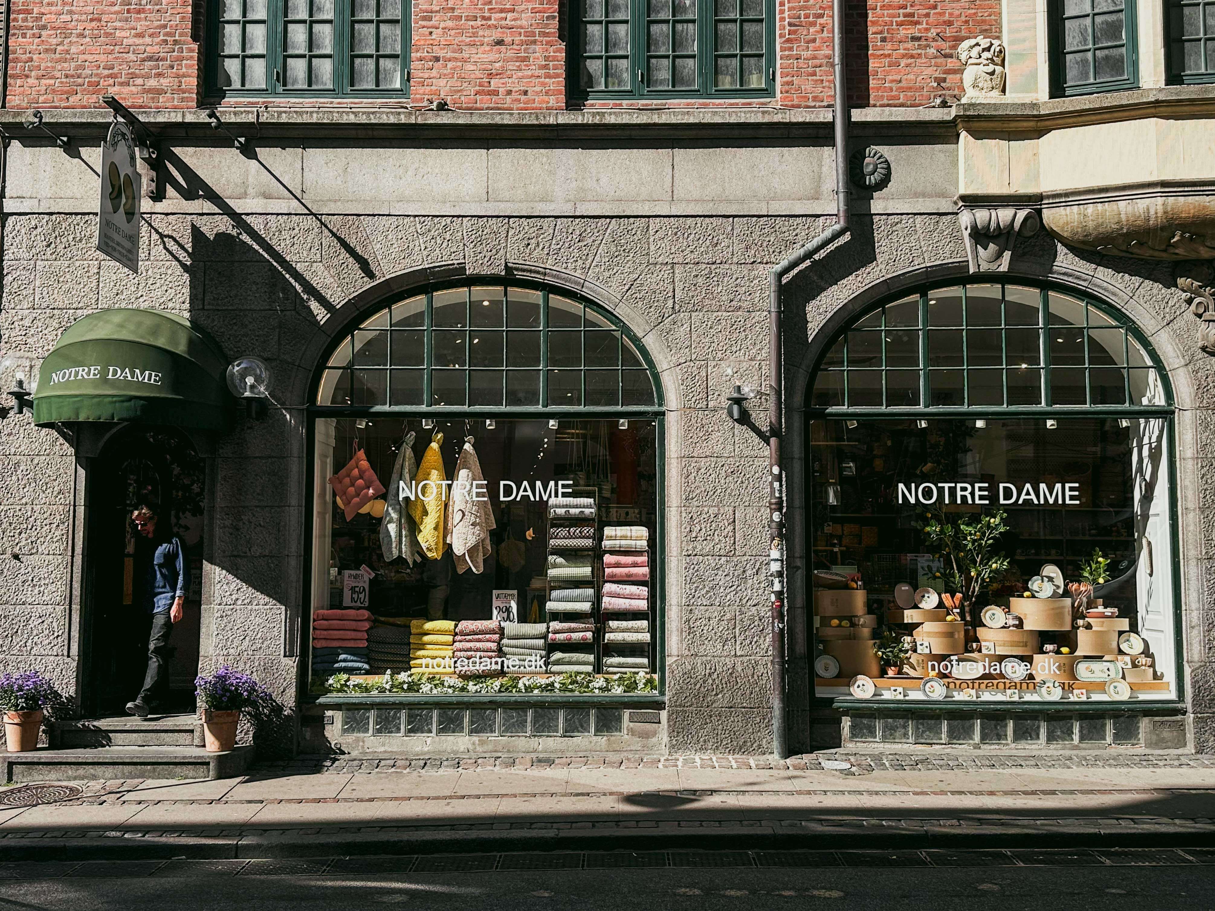 Storefront named Notre Dame with green awning, stone walls. Arched windows display colorful textiles and home goods. A person exits.
